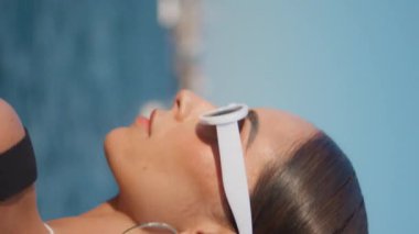 Güneşli okyanus kıyısında güneş gözlüğünü çıkaran zarif bir kadının portresi. Yaz plajında dikey olarak poz veren güzel genç bir kadın. Rıhtımda duran lüks model.