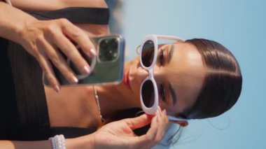 Güzel bayan cep telefonundan sosyal medyayı izliyor. Duvar kenarında dikey çekimde. Moda güneş gözlüklü mutlu, rahat kadın gülümseyerek akıllı telefon ekranına bakıyor. Zarif kız sahilde mesaj okuyor.