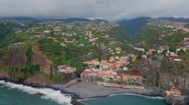 Hava yeşili deniz kenarı köyü bulutlu bir günde. Madeira adasında geleneksel evler. Kırmızı fayanslı mimarisi ve verimli manzarası olan insansız hava aracı kıyı yerleşimi. Okyanus dalgaları kayalık kıyıları yıkıyor.