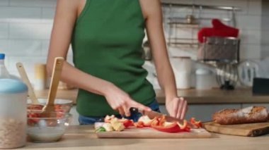 Ev mutfağında vejetaryen salata için sebze doğrayan kapanış kızı. Mutfakta kırmızı biber hazırlayan, kablosuz kulaklıkla müzik dinleyen mutlu kadın. Kulaklıklı gülümseyen bayan kahvaltı hazırlıyor..