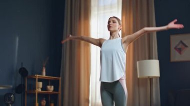 Huzurlu bir kadın evde yoga egzersizi yapıyor. Beyaz bluzlu, odaklanmış esmer, oturma odasında kollarını açmış duruyor. Sakin bayan namaste pozisyonunu alıyor. İçerde meditasyona başlıyor.