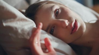 Uyanan kadın gözlerini açıyor. Yatakta güneşli bir dairede yatıyor. Yakın plan. Yatak odasındaki rahat yastıklarda uyanan nazik, güzel bir kadın. Huzurlu kız sabahın erken saatlerinde kameraya gülümsüyor. Tembel dinlenme konsepti.