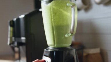 Bilinmeyen bir kadın apartmanın mutfağında smoothie hazırlıyor. Kadın eli blenderi kapatıyor ve taze yeşil vejetaryen içeceği hazırlıyor. Bayan sabahları modern mutfakta sebze karışımı pişiriyor..