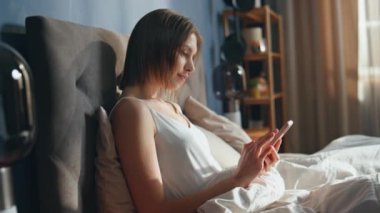 Uyanmış kız sabahın erken saatlerinde yatakta akıllı telefon kaydırıyor. Rahat bir kadın, rahat yatak odasındaki beyaz yatağa uzanmış cep telefonuyla sosyal medyada geziniyor. Tembel evde hafta sonu cep telefonu kullanan gülümseyen sakin bayan..