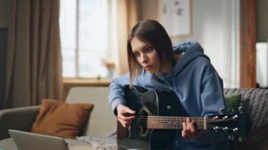 Gitar çalan yetenekli bir kadın dizüstü bilgisayarla eve yakın çekim yaparak eğitim videosu izliyor. Gülümseyen kız akustik enstrüman üzerinde melodi çalışıyor. Bilgisayarda çevrimiçi eğitime bak. Müzisyen müzik becerilerini geliştirir