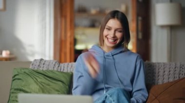 Modern apartmandaki video konferansında bilgisayarda selamlaşan kız. İnternetten konuşan gülümseyen bir kadın ev kanepede rahat rahat oturuyor. Mutlu bayan bilgisayar bağlantısıyla keyifli sohbetler.