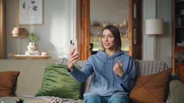 Akıllı telefondan koltukta oturan bir kadının videosu. Oturma odasında cep telefonuyla konuşan gülümseyen kadın. Arkadaş canlısı kız cep telefonuyla çevrimiçi sohbet ediyor..