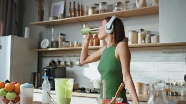 Mutfakta yeşil smoothie içen bir bayan kulaklıkla müzik dinliyor. Mutfakta besleyici vejetaryen içeceği içen mutlu kadın dans ediyor. Ev yapımı sebzelerin tadını çıkaran kız..
