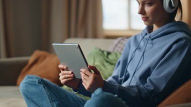 Kulaklık takmış bir kadın rahat koltukta tablet içeriği videosu izliyor. Kablosuz kulaklık takan gülümseyen kız bilgisayardaki sosyal medyada geziniyor. Rahat genç görünümlü alet ekranlı ev