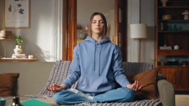 Evde meditasyon yapan kız nilüfer çiçekli koltukta rahat poz veriyor. Kapalı gözlü, namaste elli kadın sakin görünmenin tadını çıkarıyor. Rahatlamış kadın, sağlığa ve sükunete odaklanıyor..