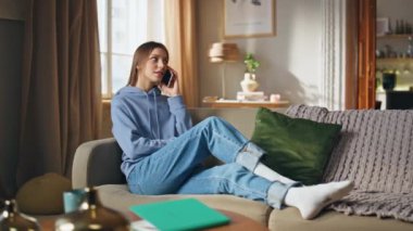 Oturma odasındaki kanepede cep telefonundan konuşan içerikli kız. Mutlu kadın cep telefonu iletişiminin keyfini çıkarıyor. Rahat bayan, modern dairede rahatlamak için akıllı telefondan arıyor..