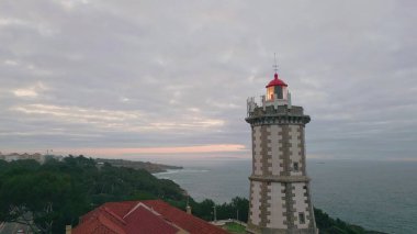 Okyanus ufkuna bakan deniz feneri bulutlu gökyüzü manzarasının altında. Modern deniz feneri yön bulmak için güzel kıyı şeridinde yanıp sönüyor. Koyu deniz suyu, dalgalı yeşil deniz kıyılarını kasvetli bir günde yıkıyor.