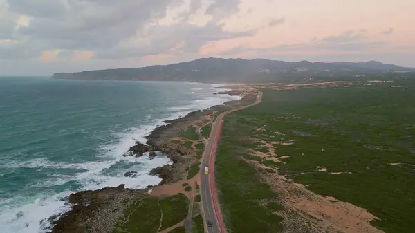 stock image Coastal roadway stretching along beautiful rough coastline aerial view. Asphalt road placed on picturesque rugged coast washed by stormy ocean waves in super slow motion. Transport riding on highway.