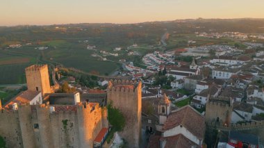 İnsansız hava aracı, gün batımıyla aydınlanan Portekiz 'in pitoresk köyündeki antik kaleyi görüyor. Ortaçağ Obidos şatosu manzaralı eski şehir çatılarının üzerinde yükseliyor. Güzel tarihi yerleşim yeri için akşam.