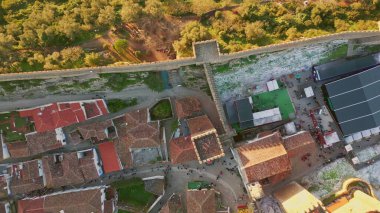 Kırmızı fayanslı, hava manzaralı köy çatıları güneşli bir akşam. Güneş ışığında eski şehri doğadan ayıran taş kale duvarı. İnsanlar geleneksel binaların etrafında tarihi caddelerde yürüyor