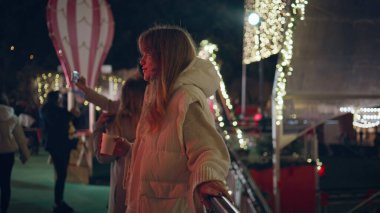 Gece festival fuarında elinde sıcak içecekle rahatlayan bir kız. Aydınlatılmış Noel ışıklarıyla çevrili karanlık sokakta duran rahat kadın. İyi tatiller. Aydınlatılmış parkta güzel bir hafta sonu..