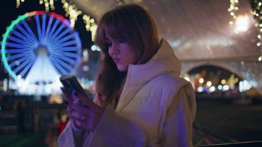 Aydınlatılmış Ay Parkı 'nda cep telefonuna bakan bir kadın. Akıllı telefon tutan mutlu kadın lunaparkta şenlik akşamının tadını çıkarıyor. Parlak ışıkların altında, sokakta mesaj atan rahat bir kız..