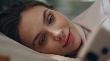 Yakın plan yalan modeli. Güneşli bir sabah uyanırken cep telefonundan gelen mesajı okuyor. Romantik gülümseyen kadın yatakta dinlenen akıllı telefon ekranına bakıyor. Güzel tembel bayan rahat yatak odasında tek başına uyuyor. 