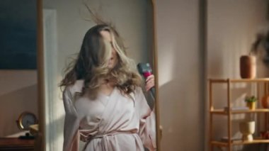 Heyecanlı ev hanımı yatak odasında saçlarını kurutup ayna yansıması yapıyor. Oturma odasında saç kurutma makinesi kullanan mutlu, güler yüzlü bayan. Bornozlu neşeli kadın duştan sonra şarkı söylüyor. 