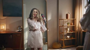 Rahat oturma odasında saç kurutma makinesi kullanan tasasız bir kız. Kurutucuda mikrofon gibi şarkı söyleyen mutlu bir kadın evde Chevelure kurutuyor. Model ayna yansıması dans ederek sabah rutini yapıyor.