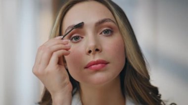 Portre yapan kadın evde kaş şekillendirici kamera yapıyor. Model kız, apartman dairesinde poz vermenin keyfini çıkarıyor. Gülümseyen kadın kaşlarını tarıyor makyaj masası görüntüsü hazırlıyor