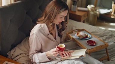 Sakin kız kahvaltıdan zevk alıyor. Yumuşak yatakta kahve içiyor. Rahat kadın sayfaları çeviriyor ve sıcak yatak odasının içinde espresso bardağı tutuyor. Güzel, gülümseyen kadın çayından bir yudum alıyor.