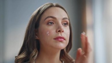 Kapalı alanda krem sürerek nemlendirici yüzü olan kadın. Güzellik uygulayan rahat bir kadın sabah cilt ürünlerini nemlendirir. Gülümseyen bir model evde aynaya bakıp şımartıyor. Kadınsı günlük prosedürler 