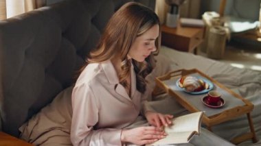 Yatakta kitap okuyan rahat bir kadın. Yakın plan. Evde kahvaltının tadını çıkaran mutlu ve çekici bir kız. Dertsiz, sırıtan kadın romanları. Yatak odasında tek başına ders çalışıyor. Sabah yaşam tarzı konsepti 