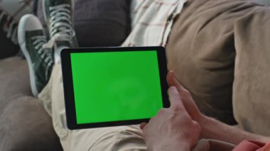 Mockup Tablet 'ten alışveriş yapan bir adam. Evdeki kanepeye yakın oturuyor. Kimliği belirsiz, yeşil ekranlı, uzaktan kumandayı seçen bir bilgisayar. Krom anahtar aygıtı kullanarak proje üzerinde çalışan serbest çalışan bir erkek..