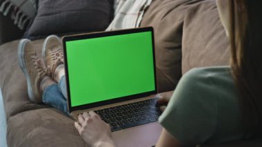 Yeşil ekran dizüstü bilgisayarda Webinar izleyen bir kadın. Evdeki rahat kanepeye yakın oturuyor. Bilinmeyen bir kadın dairedeki koltukta krom anahtar bilgisayarına bakıyor. Uzaktan kumandalı dijital aygıt kullanan serbest çalışan bir kadın.