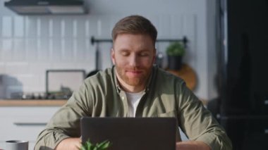 Gülümseyen adam videosu ev mutfağından dizüstü bilgisayarı arıyor. Happy Freelancer, modern mutfaklarda oturup bilgisayar sohbeti yaparak iş görüşmesi yapıyor. İçerideki konferansta belgelere bakan olumlu adam.
