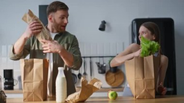 Apartman mutfağında paketleri boşaltan gülen eşler yakın planda takılırlar. Mutlu erkek kadın yemekle kavga ediyor evde eğleniyor. Modern mutfaktaki ev işleri üzerine neşeli bir çift bağ..