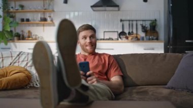 Mutlu adam cep telefonu okuyup rahat koltukta oturuyor. Akıllı telefondan gülümseyen erkek mesajları oturma odasında rahat bir hafta sonu geçiriyor. Konforlu apartman kanepesinde sohbet eden olumlu adam..