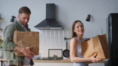 Aile çifti, beyaz mutfak iç kısımlarında yiyecek paketlerini boşaltıyorlar. Sağlıklı yiyeceklerle dolu kağıt torbaları eve götüren mutlu bir adam ve kadın. Modern dairede ev işlerini paylaşan sevimli bir çift..