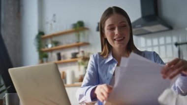 Ev mutfağında bilgisayarla konuşan gülümseyen öğretmen. Pozitif kadın öğretmen video konferansında eğitim planını tartışıyor. İnternet konferansında konuşan neşeli bir kadın iş belgelerini tutuyor..