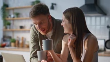 Aile bilgisayarı mutfakta kahve fincanlarını yakın çekimde tutuyor. Rahatlamış genç eşler internet üzerinden alışveriş yapıyorlar. Ev mutfağından. Sabah içkilerinin keyfini çıkarın.