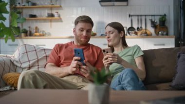 Mutlu çift koltukta dinleniyor, oturma odasında cep telefonlarına bakıyor. Gülümseyen adamla akıllı telefondan online alışveriş hakkında konuşan gülen kadın. Mutlu eşler evde cep telefonlarını tutarak sohbet ediyorlar..