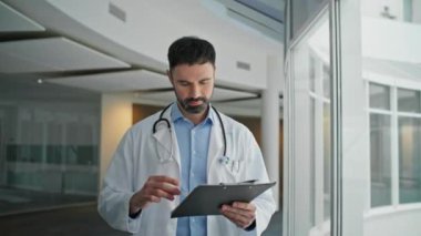 Doctor reviewing patient information on clipboard. Closeup smiling man walking checking test results modern clinic. Healthcare professional examining chart going to consultation. Happy bearded expert.