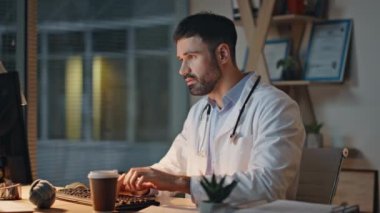 Doktor gece vardiyası sırasında tıbbi belgeleri kontrol ediyor. Kapalı, yorgun sıhhiyeci akşam hastanesinde uykulu hissederek kahve içiyor. Klinikte steteskopla klavye yazan odaklanmış bir doktor.