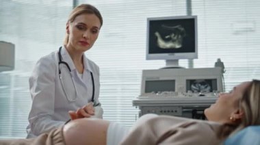 Kadın klinikte ultrason çekiyor. Doğum kontrolü sırasında profesyonel jinekologla konuşmaktan mutluluk duyarız. Gülümseyen doktor tarama makinesi kullanıyor. Modern doğum öncesi muayene.