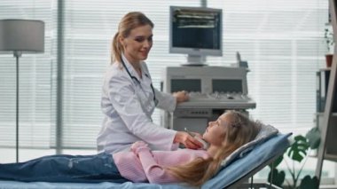 Profesyonel ultrason muayenesi koğuşta. Gülümseyen doktor salgı bezini kontrol ediyor ve genç hastayla koltukta konuşuyor. Modern klinikte sonografi uzmanı kontrol prosedürü uyguluyor