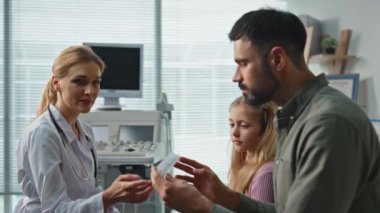 Kadın doktor, çocuğun babasıyla ilaçları tartışıyor. Klinik randevusu sırasında ilaç kutusunu inceleyen yakın bir ebeveyn. Arkadaşça gülümseyen terapist modern sağlık merkezindeki aileye danışmanlık yapıyor..