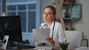 Doktor internetten hasta belgelerini inceliyor. Üniformalı, gülümseyen bir kadın akşam kliniğinde test sonuçlarını tartışıyor. Gülümseyen sağlık uzmanı tıbbi kayıtları analiz eden belgeleri tutuyor.