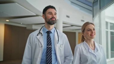 Gülümseyen doktorlar modern klinikte birlikte yürürler. Kapalı alan sağlık uzmanları içeride olumlu ve kararlı olduklarını gösteriyorlar. Tıbbi uzmanlar koridora bakan kameralara bakıyorlar. Doktor ekibi hastanede.