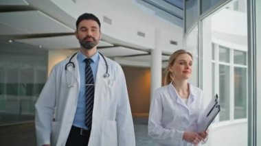 Klinik çalışanları birlikte yürüyorlar. Yakın plan. Sağlık merkezinde çalışan iki doktor. Steteskoplu, kendine güvenen sakallı adam iş arkadaşıyla hastaneye gidiyor. Profesyonel kadın, pano taşıyor. 