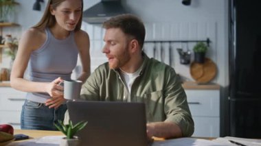 Serbest çalışan, ev mutfağında laptopta çalışan karısından kahve alıyor. Gülümseyen kadın mutfaktaki meşgul kocaya bardak içeceği getiriyor. Sakallı adam, kadın bakımı ve desteği için minnettar. Aile sevgisi