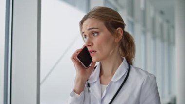 Hastanede akıllı telefondan konuşan endişeli bir pratisyen. Stresli doktor şakaklarına masaj yapıyor ve sağlık teşhisini tartışıyor. Beyaz önlüklü, üzgün bir sağlık uzmanı. Endişelerini ve ciddiyetlerini dile getiriyor.