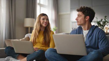 Kanepe kapanışında dizüstü bilgisayarları karıştıran neşeli sevgililer. Mutlu adam pozitif kadın klavyesini yazarken gülümseyerek birbirine bakıyor. Apartman dairesinde birlikte çalışan genç çift.