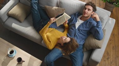 Genç eşler rahat kanepe manzarasında kitap okuyor. Gülümseyen, rahat bir çift, sakin bir ortamda samimi bir anın tadını çıkarıyor. Neşeli adam eşiyle konuşuyor. Oturma odasında birlikte rüya görüyorlar.