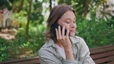 Akıllı telefondan bahseden gülümseyen bir kadın Serene Park 'ta yakın planda oturuyor. Etrafı yeşilliklerle çevrili, huzurlu sohbetin keyfini çıkaran mutlu kız. Rahat esmer, güzel yeşil bahçedeki bankta cep telefonunu arıyor.